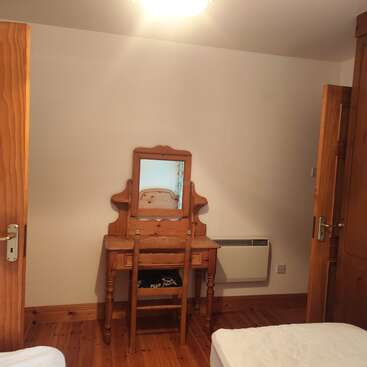 Dieses Bild zeigt ein schlichtes Schlafzimmer mit Holzmöbeln, darunter ein Schminktisch, ein Spiegel und ein Stuhl. Es gibt zwei Türen, einen Holzboden und schlichte Wände.