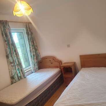 Das Bild zeigt ein schlichtes Schlafzimmer mit zwei Einzelbetten, einem Nachttisch aus Holz, geblümten Vorhängen, einem Holzboden, weißen Wänden und einer gelben Deckenlampe.