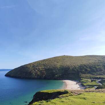 Eine atemberaubende Küstenlandschaft mit einem grünen Hügel, der einen ruhigen, halbmondförmigen Sandstrand überragt, klarem, blauem Meerwasser und einem strahlend blauen, wolkenlosen Himmel darüber.