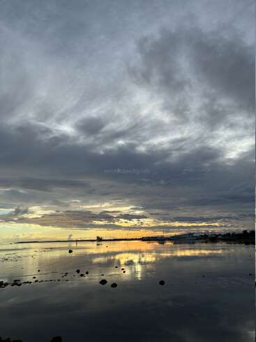 A dramatic sky filled with clouds reflects beautifully on the calm waters below, with distant boats and a peaceful harbor at sunset, creating a serene atmosphere.