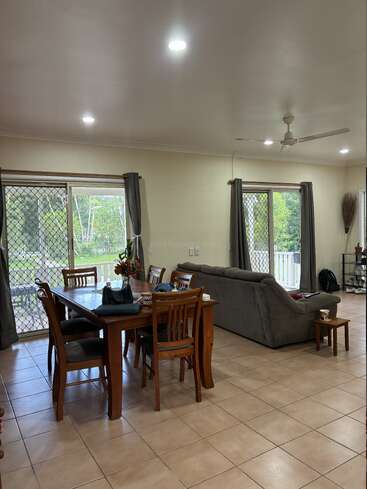 A cozy living and dining area features a wooden table with chairs, grey couch, large windows with curtains, ceiling fan, tiled floor, and natural light.