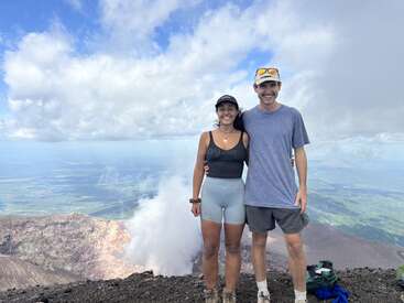 A smiling couple stands on a rocky mountain peak above the clouds, dressed in casual hiking gear. Lush landscape and dramatic sky create a breathtaking backdrop.