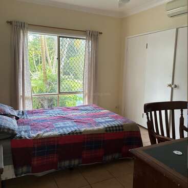 A cozy bedroom features a bed with a colorful patchwork quilt, a wooden chair, large window with garden view, white closet doors, and light curtains.