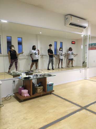 The image depicts a dance studio with five individuals standing on a raised platform, wearing masks and facing a large mirror, with a small table in front of them.