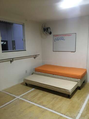 The image depicts a small room with a window, a whiteboard, and two beds, featuring a white wall, beige floor, and a metal handrail along the left wall.