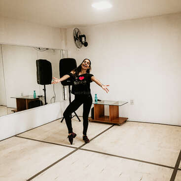 The image depicts a woman dancing in a studio with white walls, a mirror, and a speaker, wearing a black shirt and pants, with her arms outstretched.