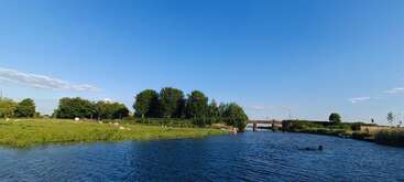 A peaceful riverside scene with calm blue water, lush green grass, trees, and a small bridge under a clear, bright blue sky, exuding tranquility.