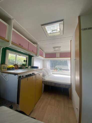 This image shows the inside of a cozy camper with pastel cabinets, a small kitchenette, a bed by the window, wood flooring, and natural light.