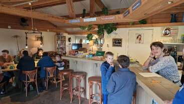 A cozy café interior with wooden beams and warm lighting. People sit at tables and the bar, chatting, while plants and books add a homely touch.