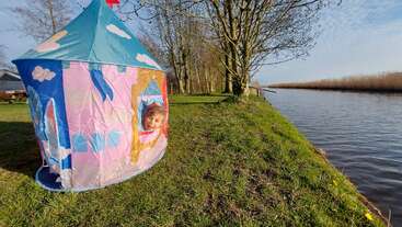 A young child smiles from the window of a colorful castle-shaped play tent set on green grass near a peaceful river, surrounded by trees under blue sky.