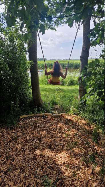 A girl with long hair sits on a swing between two trees, overlooking a green field and river, surrounded by lush foliage on a sunny day.
