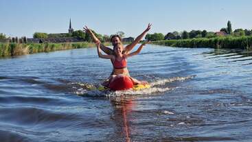 Two people are riding a red inflatable towable on a calm river, smiling with arms raised, surrounded by green fields and trees under a clear blue sky.