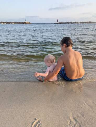 Ein kleiner Junge und ein Baby sitzen zusammen am Ufer und genießen an diesem ruhigen Strandtag das ruhige Wasser und die warmen Sonnenstrahlen.