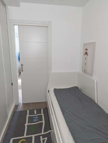 Ein kleiner, heller Raum mit weißen Wänden, in dem ein Einzelbett mit einer grauen Decke, ein Bild von einem Leuchtturm, ein Hopse-Teppich, ein Schrank und eine Tür stehen.