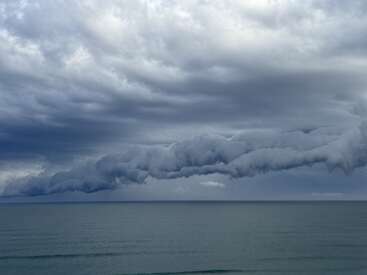 Eine dramatische, dunkle Rollwolke erstreckt sich über den Himmel über einem ruhigen Meer. Die stimmungsvolle, graue Atmosphäre deutet auf einen bevorstehenden Sturm hin, während das Wasser darunter ruhig ist.