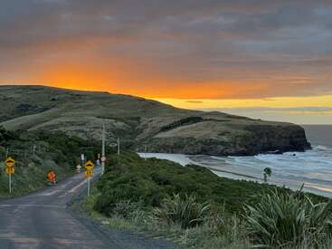 A winding coastal road leads toward a serene beach and rolling hills at sunset. Dramatic orange sky glows above the ocean, creating a tranquil, picturesque scene.