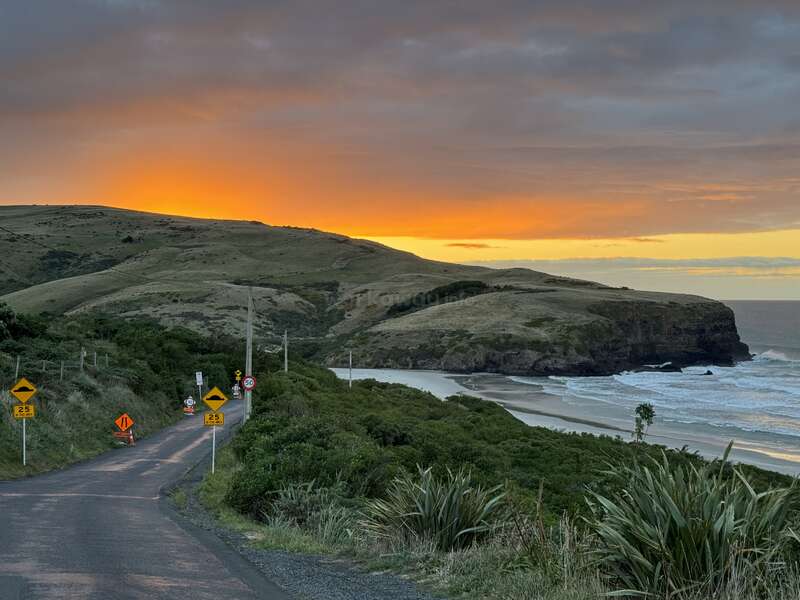Eine kurvenreiche Küstenstraße führt zu einem ruhigen Strand und sanften Hügeln bei Sonnenuntergang. Der orangefarbene Himmel leuchtet über dem Meer und schafft eine ruhige, malerische Kulisse.