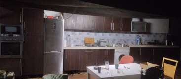 Essa imagem mostra uma cozinha meio escura com armários escuros, uma geladeira, um forno, uma máquina de lavar e uma mesa com copos, cadeiras e alguns itens espalhados.