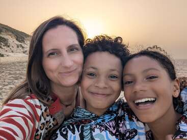 Eine Frau und zwei lächelnde Kinder posieren für ein Selfie an einem Sandstrand bei Sonnenuntergang, mit warmem Licht und Klippen im Hintergrund, die Glück ausstrahlen.