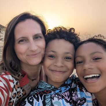 Una mujer y dos niños sonrientes posan para un selfie en una playa de arena al atardecer, con luz cálida y acantilados de fondo, irradiando felicidad.