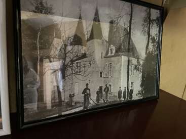Una fotografía enmarcada en blanco y negro muestra a varias personas de pie ante un edificio histórico con torres, rodeado de árboles desnudos, expuesto sobre una superficie de madera en el interior.