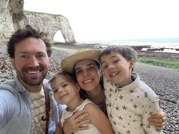 Una sonriente familia de cuatro miembros se toma un selfie en una playa rocosa con espectaculares acantilados y el mar de fondo. Se les ve felices, cercanos y relajados.