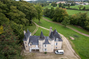 A picturesque chateau with turrets sits surrounded by lush green trees and rolling fields, accessed by winding dirt roads, with a white van parked outside.