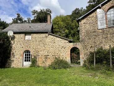 This image shows two rustic stone buildings connected by an archway, surrounded by greenery and trees, under a partly cloudy sky, with shuttered windows.