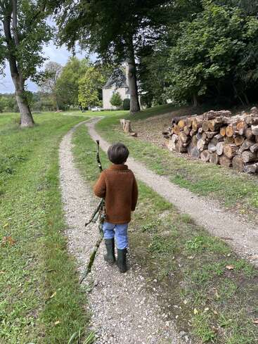 A child in a brown sweater and boots walks down a gravel path, carrying a large stick, beside stacked firewood and lush green trees.