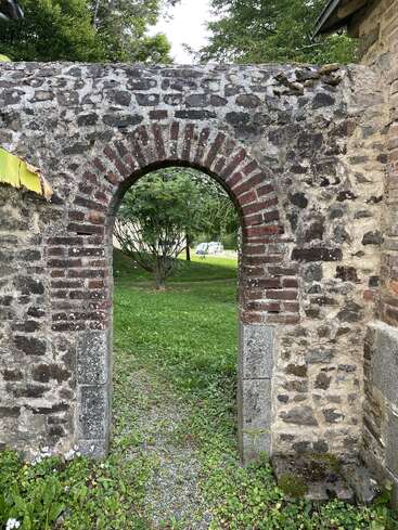 An old stone archway with red bricks forms a doorway in a rustic wall, leading to a lush green garden with trees and grass beyond.