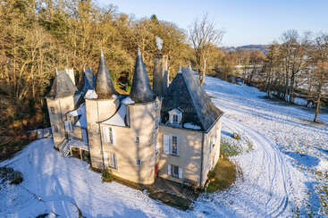 Un château pittoresque aux multiples tours pointues se dresse au milieu d'un paysage enneigé, entouré d'arbres dénudés et de champs, sous un ciel bleu limpide. De la fumée s'élève doucement.