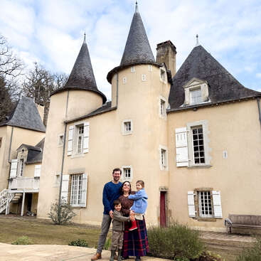 A smiling family of four stands in front of a charming, fairytale-like chateau with beige walls, pointed turrets, many windows, and a cloudy blue sky overhead.