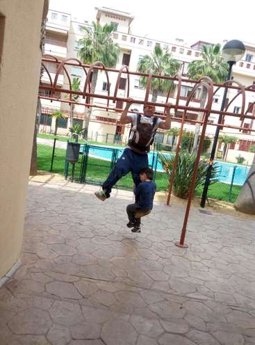 Das Bild zeigt einen Mann, der ein Baby trägt, während ein anderes Kind auf einer Spielplatzstange schaukelt, vor einem Hintergrund aus Palmen und Wohnhäusern.