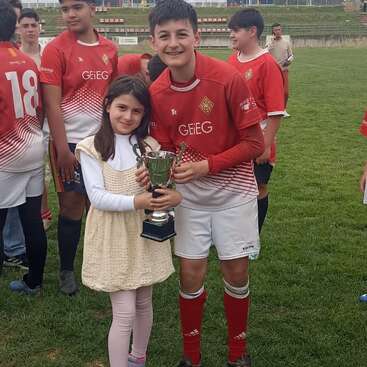 A young girl and a boy in a red soccer uniform proudly hold a trophy together on a grassy field, surrounded by their smiling teammates.