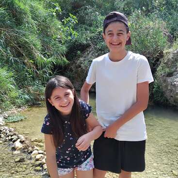 Um menino e uma menina sorridentes estão ao lado de um riacho raso cercado por vegetação e pedras, desfrutando de um dia ensolarado ao ar livre. Eles parecem felizes e brincalhões juntos.