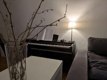 En una acogedora sala de estar hay un piano digital con partituras, una lámpara de pie encendida, un sofá gris, una mesa blanca y un jarrón de cristal con ramas secas.