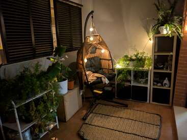 Un rincón acogedor y con luz tenue cuenta con una silla colgante con guirnaldas de luces, rodeada de exuberantes plantas verdes, estanterías con espacio de almacenamiento y una alfombra tejida en el suelo.