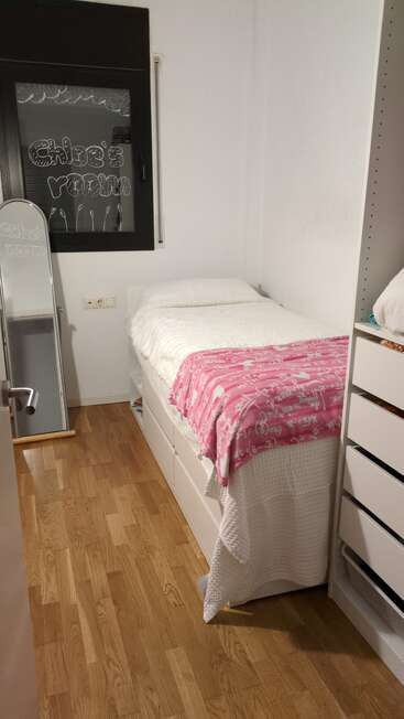Una habitación pequeña y acogedora con una cama individual, ropa de cama blanca, una manta rosa, suelo de madera, un espejo, una cómoda y una ventana en la que se lee «Habitación de Chloe».
