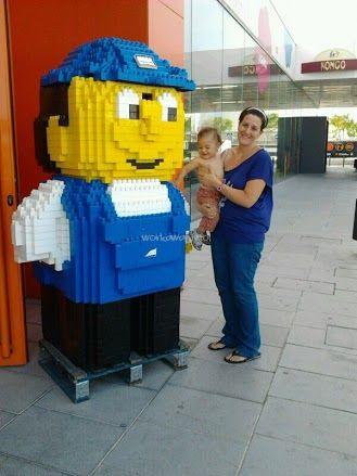 Das Bild zeigt eine Frau mit einem Baby im Arm, die neben einer großen Lego-Statue eines Postboten vor einem Glasgebäude mit einem roten Schild steht.