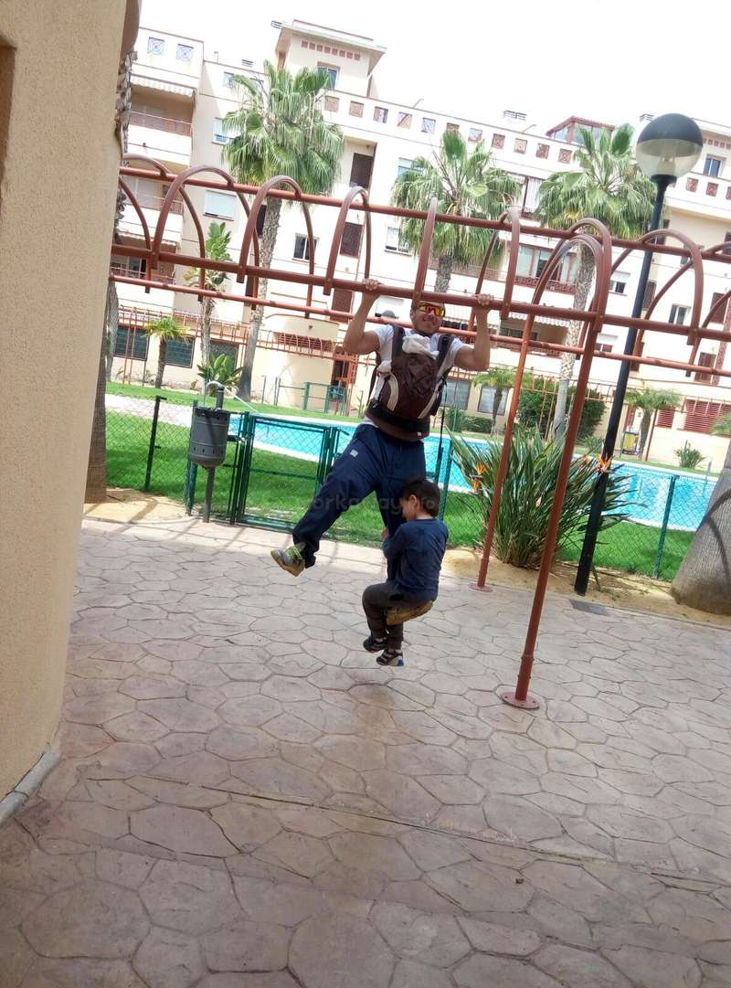 Das Bild zeigt einen Mann, der ein Baby trägt, während ein anderes Kind auf einer Spielplatzstange schaukelt, vor einem Hintergrund aus Palmen und Wohnhäusern.