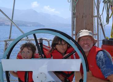 L'image montre un homme et deux enfants sur un bateau, portant des gilets de sauvetage, posant derrière un volant, avec un plan d'eau et des montagnes en arrière-plan.