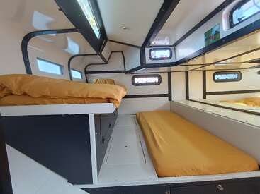 L'image représente l'intérieur d'un camping-car, avec deux lits à la literie jaune, des fenêtres et une kitchenette compacte, offrant ainsi un espace de vie confortable et fonctionnel.