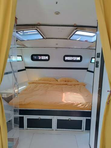 L'image représente l'intérieur d'un petit camping-car blanc avec un lit, des lucarnes et des fenêtres, avec des rideaux et une literie jaunes.