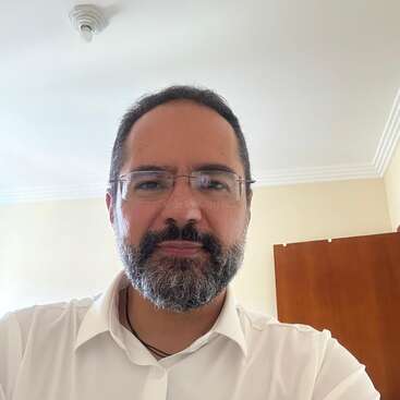 Un hombre con gafas y barba, vestido con camisa blanca, posa para un selfie en una habitación luminosa con paredes beige y muebles de madera.
