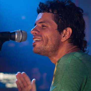 La imagen muestra a un hombre cantando frente a un micrófono, con una camisa verde y las manos levantadas, sobre un fondo azul.