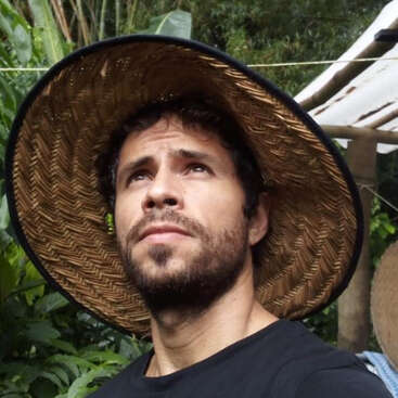 Un homme barbu portant un grand chapeau tressé à larges bords regarde vers le haut d'un air pensif. Il se tient à l'extérieur, entouré de plantes vertes et de feuillages rustiques et tropicaux.