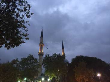 Das Bild zeigt eine Moschee mit zwei Türmen, umgeben von Bäumen und Straßenlaternen, vor einem bewölkten Himmel in der Abenddämmerung.