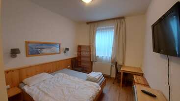 The image depicts a room with a bed, desk, and chair, featuring a bookshelf and a window. The walls are painted white, and the floor is made of wood. The room is well-lit and organized.