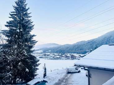 A serene winter village scene, snow covers rooftops and ground, tall pine tree left, white van parked, distant frosty hills and soft pastel sky visible.