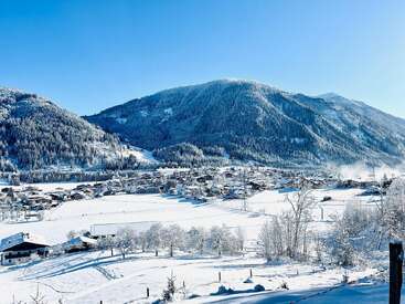 A picturesque village nestles in a snowy valley, surrounded by frosted trees and majestic mountains under a clear blue sky, creating a stunning winter landscape.